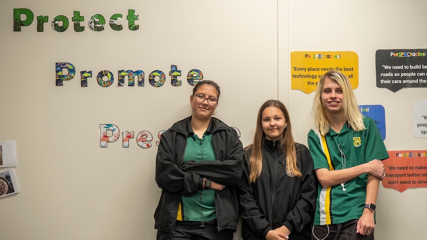Three young students lean against a wall with the words Protect, Promote behind them.