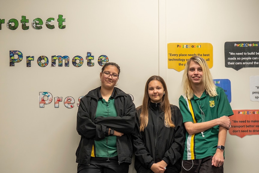 Three young students lean against a wall with the words Protect, Promote behind them.