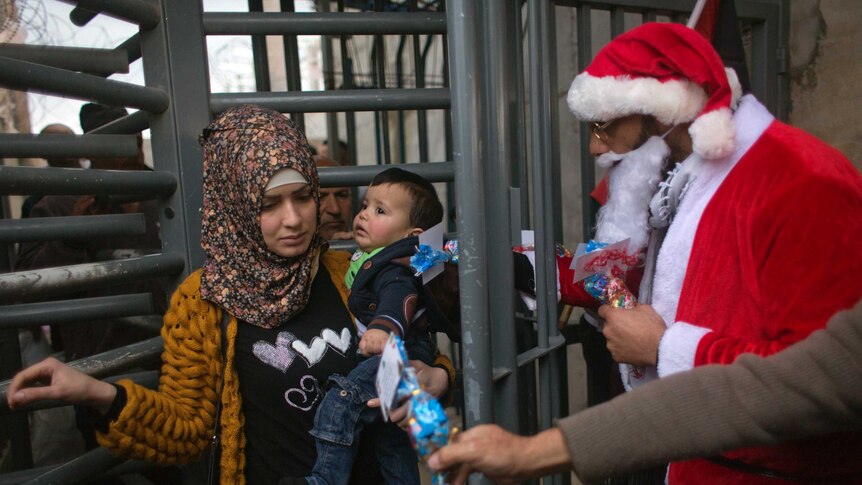 A woman carries a little boy on her hip as she walks through a barrier. One man dressed as Santa watches her another reaches out