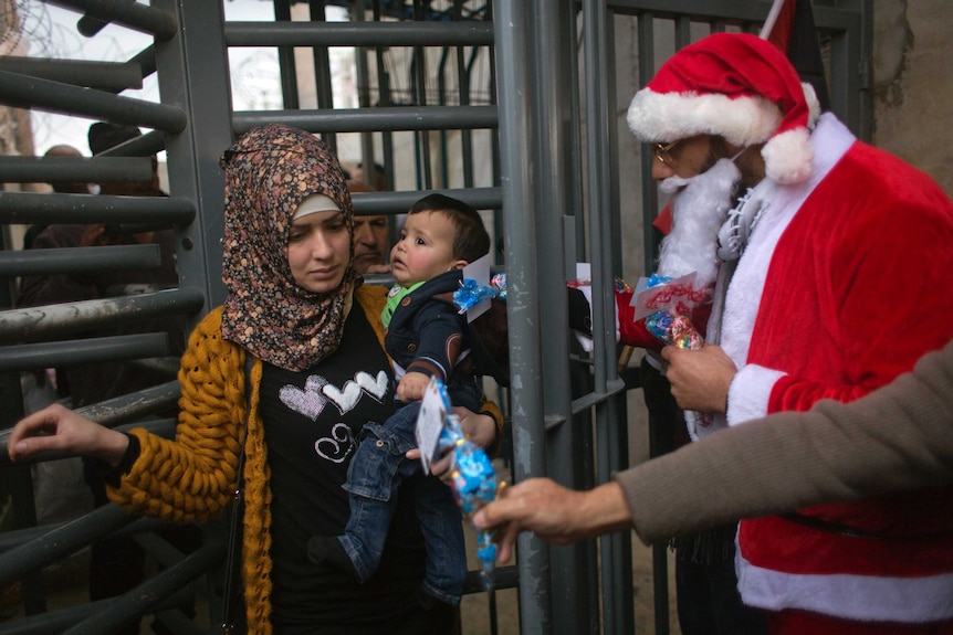 A woman carries a little boy on her hip as she walks through a barrier. One man dressed as Santa watches her another reaches out