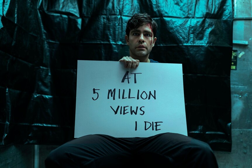 Dark haired man sits in front of black tarp looking scared and holding a sign that reads "At 5 million views I die".