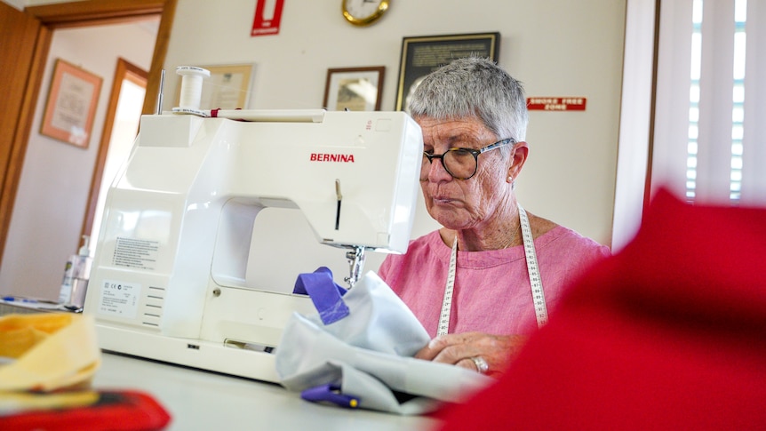 A woman wearing glasses with measuring tape around her neck peers at a sewing machine sitting on a table.