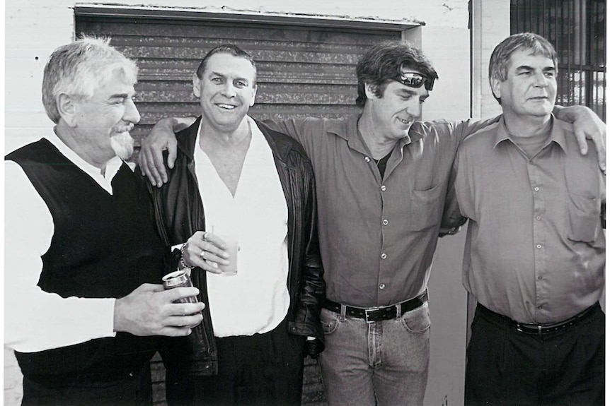 Black and white image of four middle aged men standing side by side, one with arms around others
