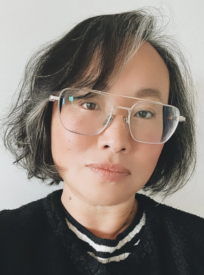 Close-up photo of person with black and white hair, large glasses and a closed-mouth smile.