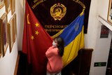 A Chinese woman in a mask and pink jumper stands between a Chinese and Ukrainian flag.