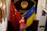 A Chinese woman in a mask and pink jumper stands between a Chinese and Ukrainian flag.