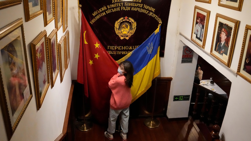 A Chinese woman in a mask and pink jumper stands between a Chinese and Ukrainian flag.