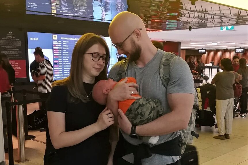 Belma and Demir Cancar hold baby Zeid in front of a departure sign at an airport