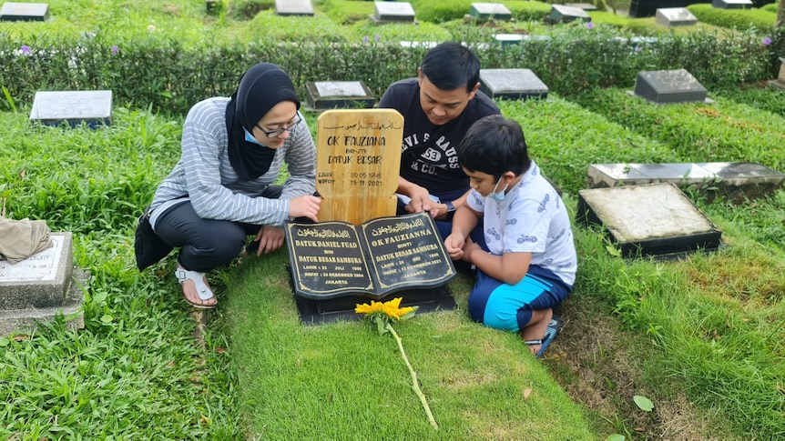 Three people sit at a grave site with their heads down.
