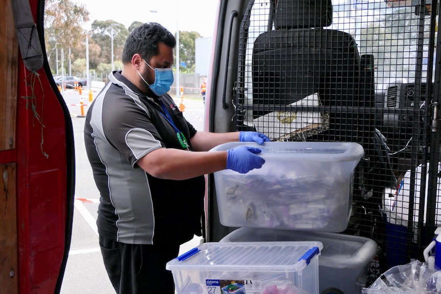 A man in a surgical mask and gloves loads containers of COVID swabs into a van