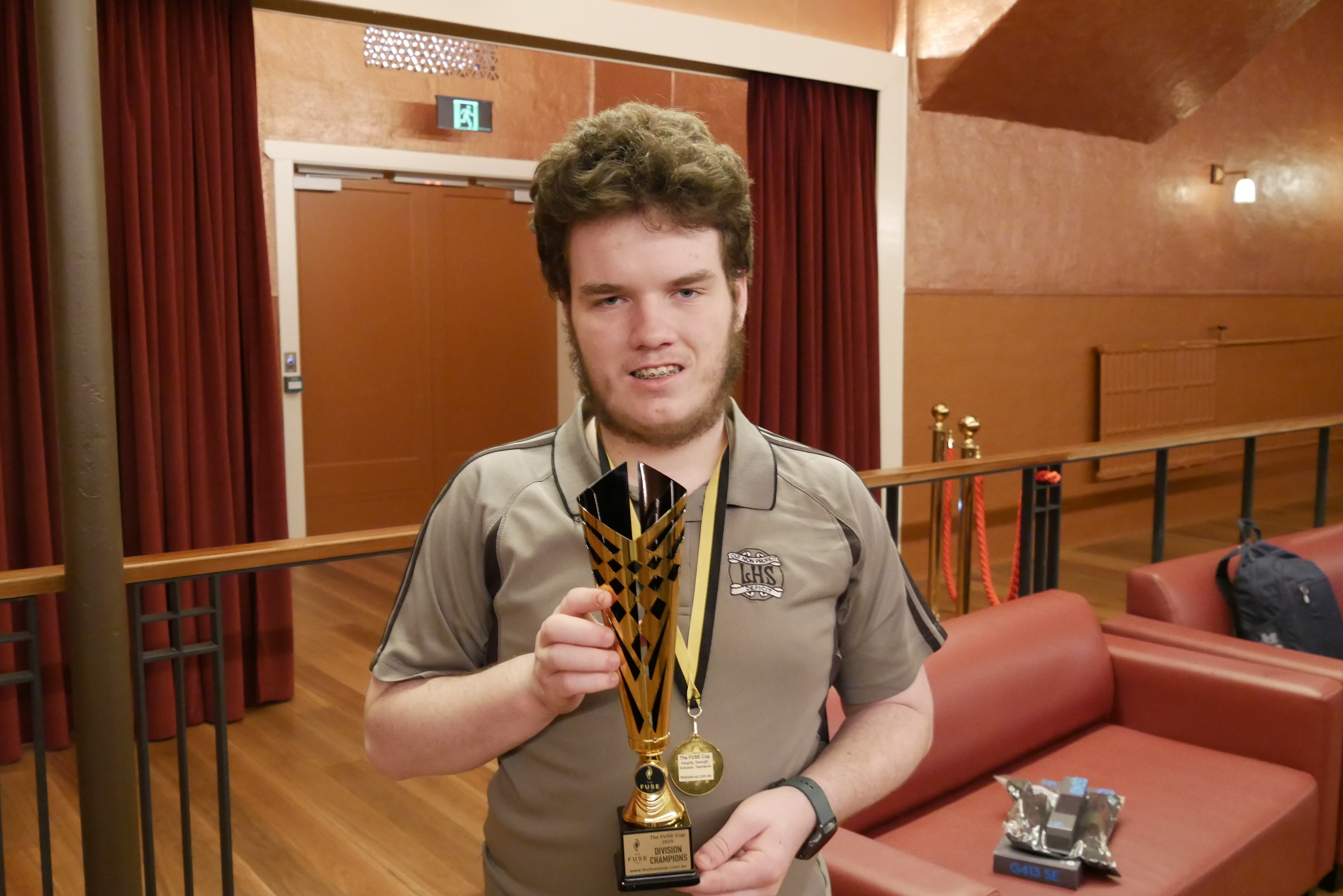 Teen boy with brown hair and beard stands in a room holding a trophy and wearing a medal around his neck 