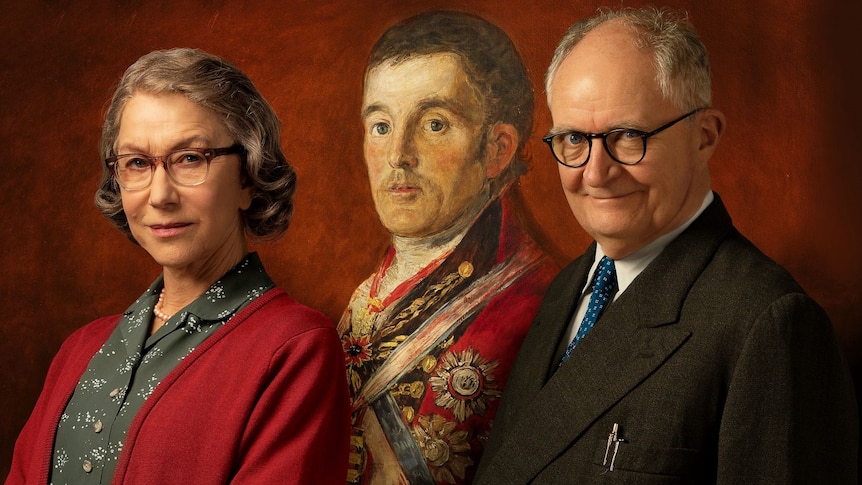 Grey-hair white woman wears red cardigan and glasses and stands beside balding man with glasses and black suit.