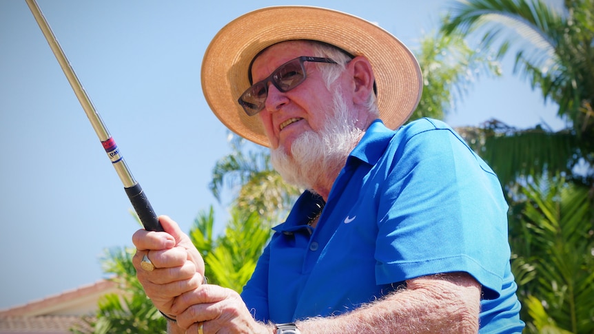 Elderly man standing outside holds a golf club high and looks out of shot.