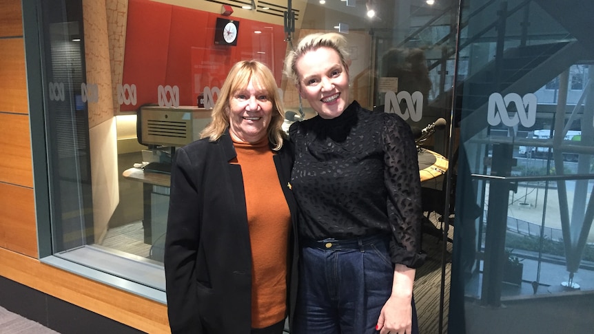 two women standing outside a glass walled ABC radio studio, smiling.