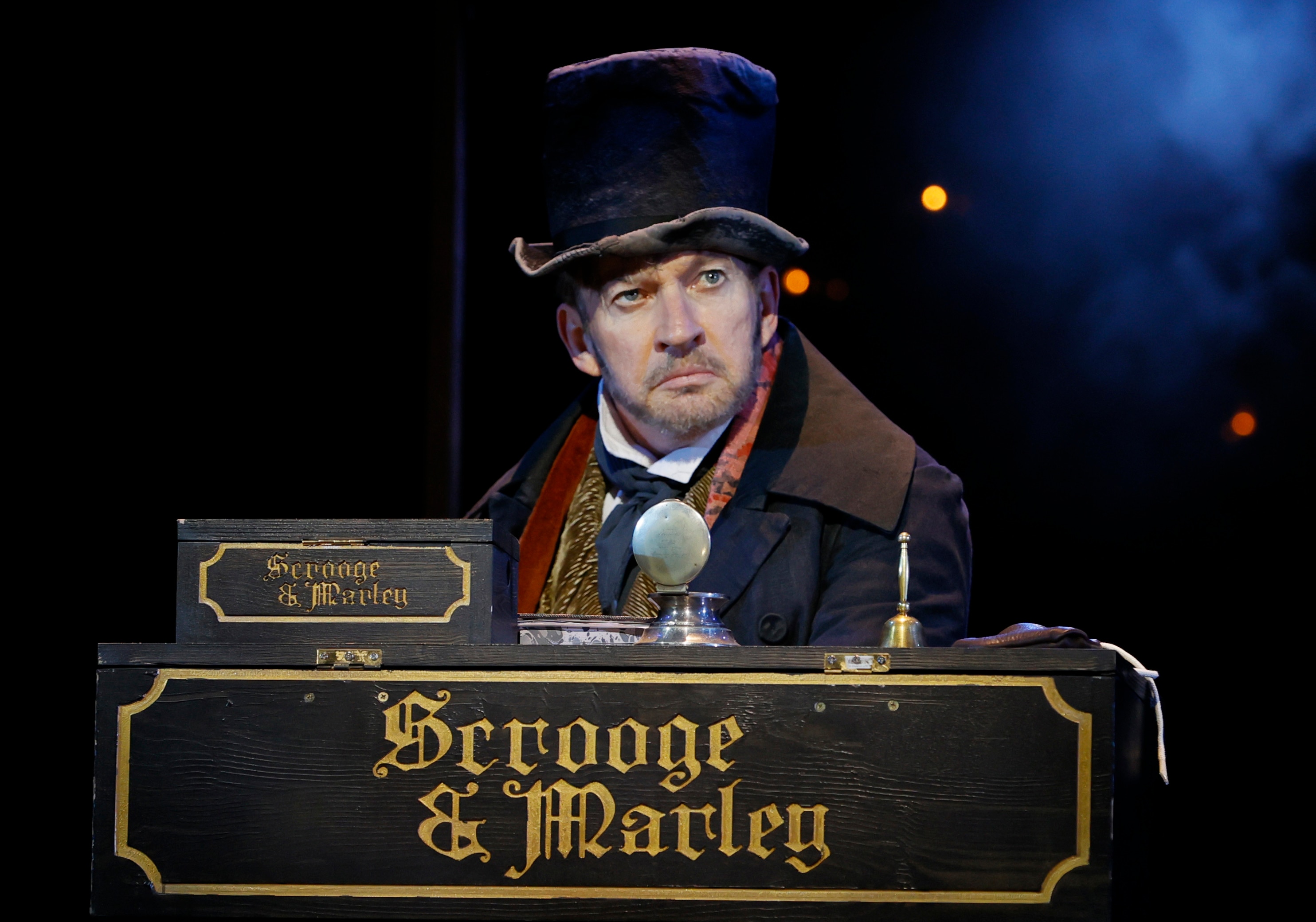 On stage, a middle-aged man in Victorian era dress looks disgruntled, standing behind a desk that reads "Scrooge & Marley"