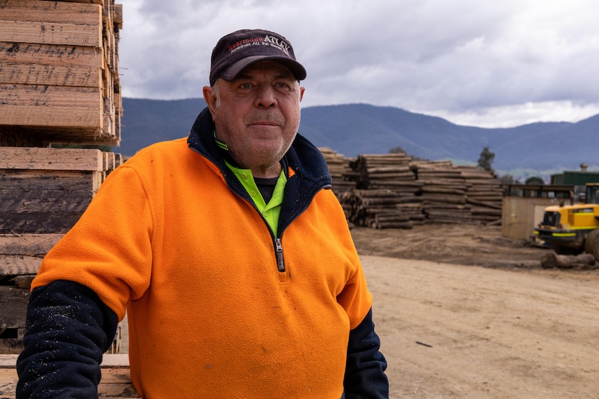 A man in a high-vis jumper stands next to pallets with logs piled in the background.