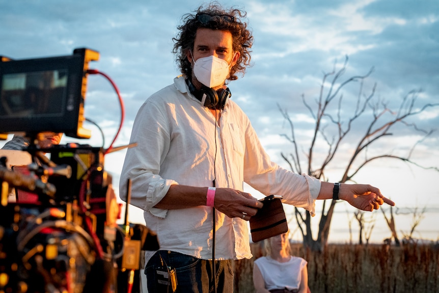 man with curly hair, wearing a face mask, standing beside filming equipped with dead gums in the backdrop