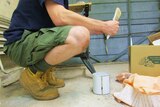 A man in work boots holding a paint brush knees down to a paint tin.