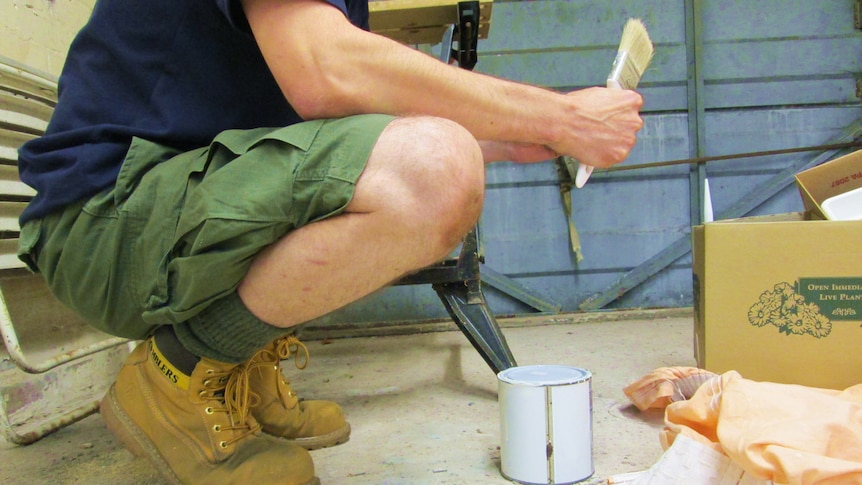 A man in work boots holding a paint brush knees down to a paint tin.