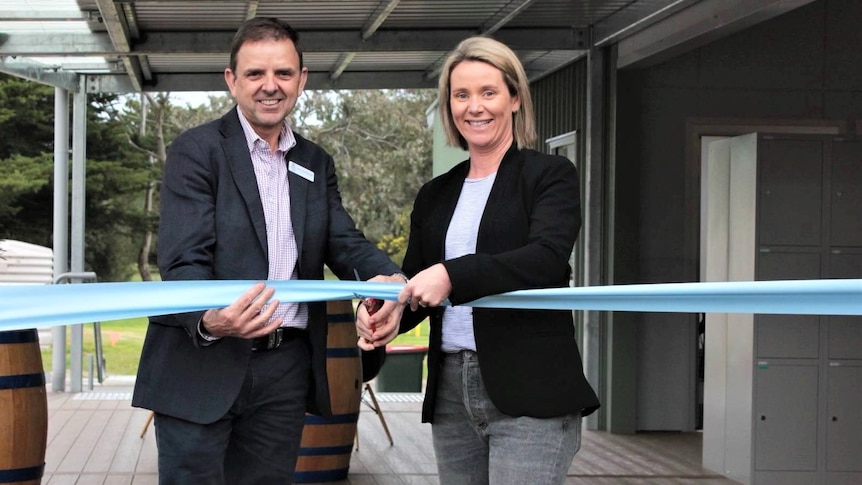 A man and a woman grin as they cut a big blue ribbon in front a school building
