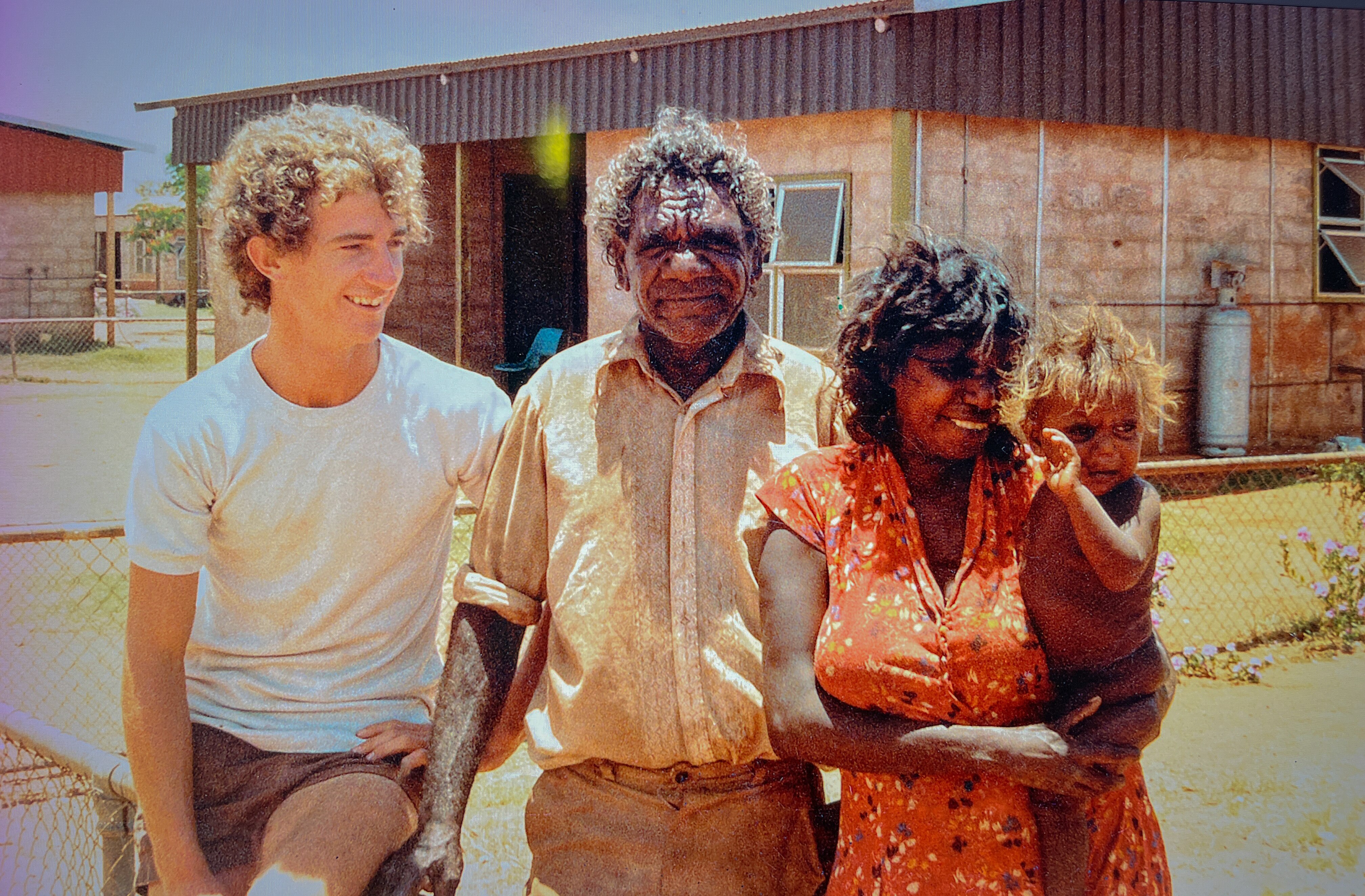 Una mujer con un niño pequeño en la cadera sonríe junto a otros dos hombres frente a un edificio de ladrillos.