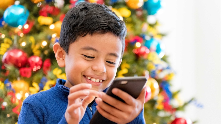 A boy stands before a Christmas tree with a smartphone in his hands