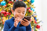 A boy stands before a Christmas tree with a smartphone in his hands