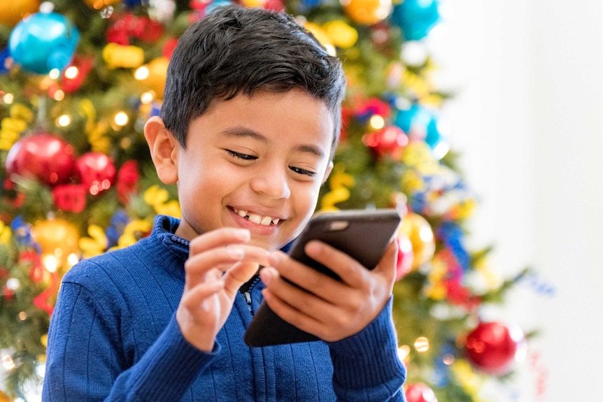 A boy stands before a Christmas tree with a smartphone in his hands