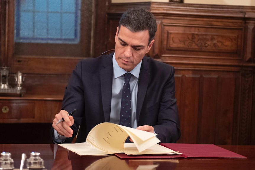 Spanish Prime Minister Pedro Sanchez signs document on mahogany desk in room with wood pannelling.