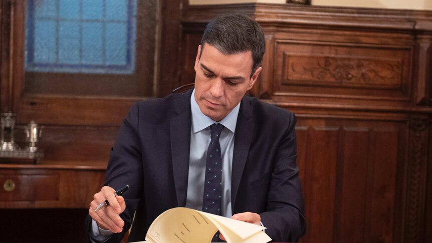 Spanish Prime Minister Pedro Sanchez signs document on mahogany desk in room with wood pannelling.