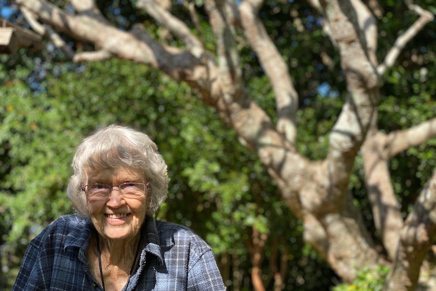 An elderly woman stands in the mottled shade of her garden with a very big frangipani trunk in the background