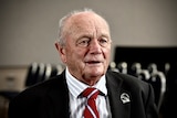 An elderly man wearing a striped blue shirt, striped red tie and black jacket is mid-sentence with rows of chairs behind him.