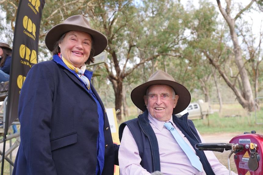 A woman wearing a hat and blue blazer smiles next to a man in a mobility scooter wearing a hat