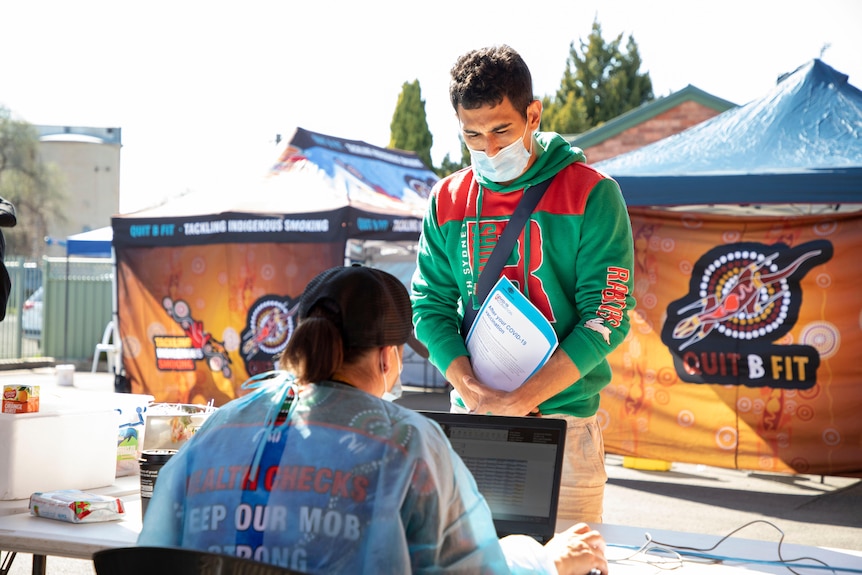 A young man holding a COVID vaccination brochure speaks to a worker in full PPE at a desk.