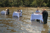 White tablecloths drape over six tables that stand in a row in the river with two people at each