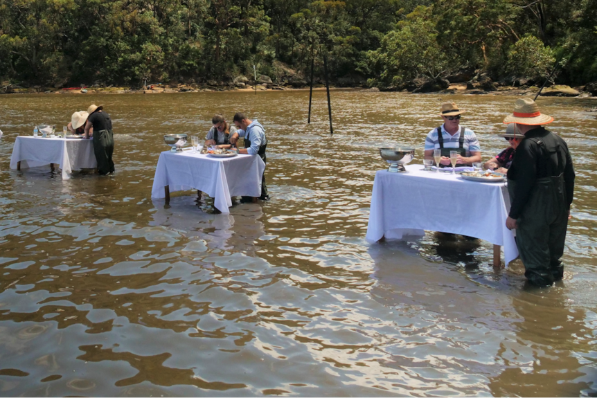 White tablecloths drape over six tables that stand in a row in the river with two people at each