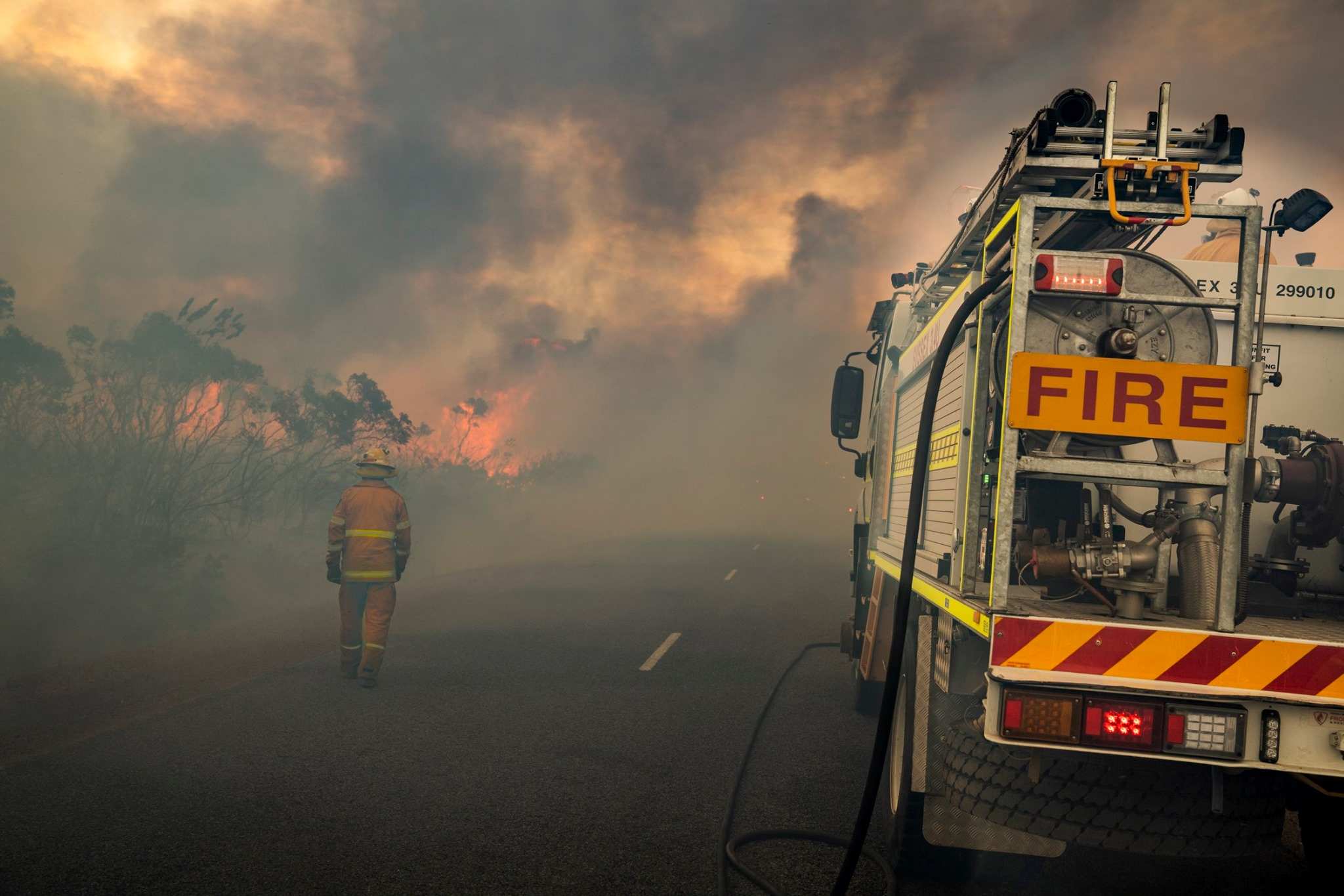 A firefighter walks along a road with flames in front of him. Smoke is everywhere.