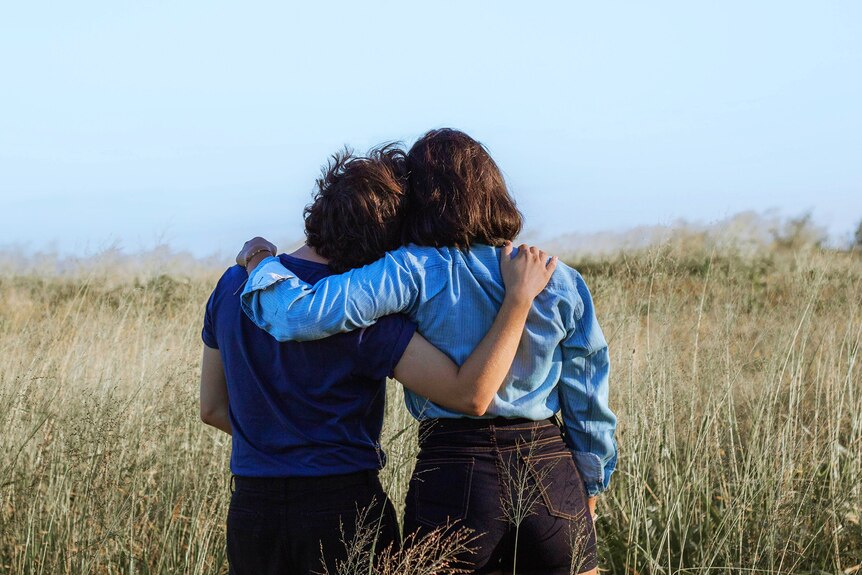 Two female friends stand with their arms around each other, looking across a field