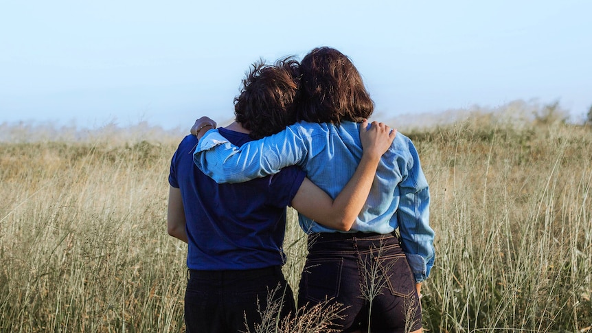 Two female friends stand with their arms around each other, looking across a field