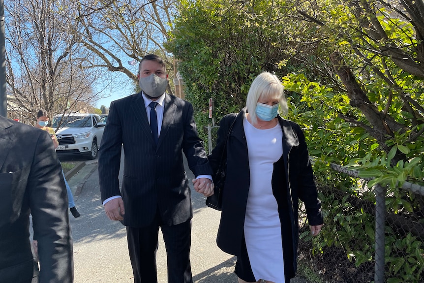 A man wearing a suit and tie walks beside an older woman wearing a black and white dress, holding her hand