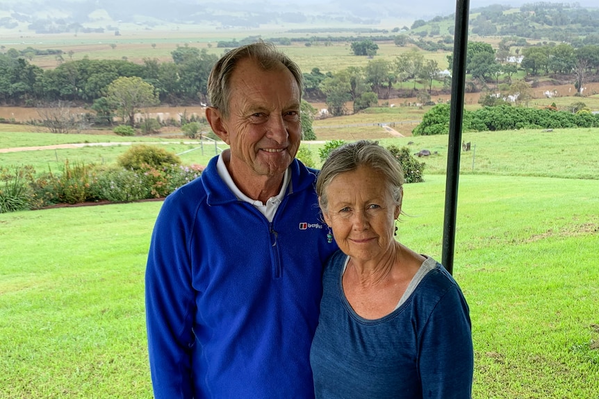 A man and woman wearing blue shirts stand on a verandah on their farm with a river in the background.