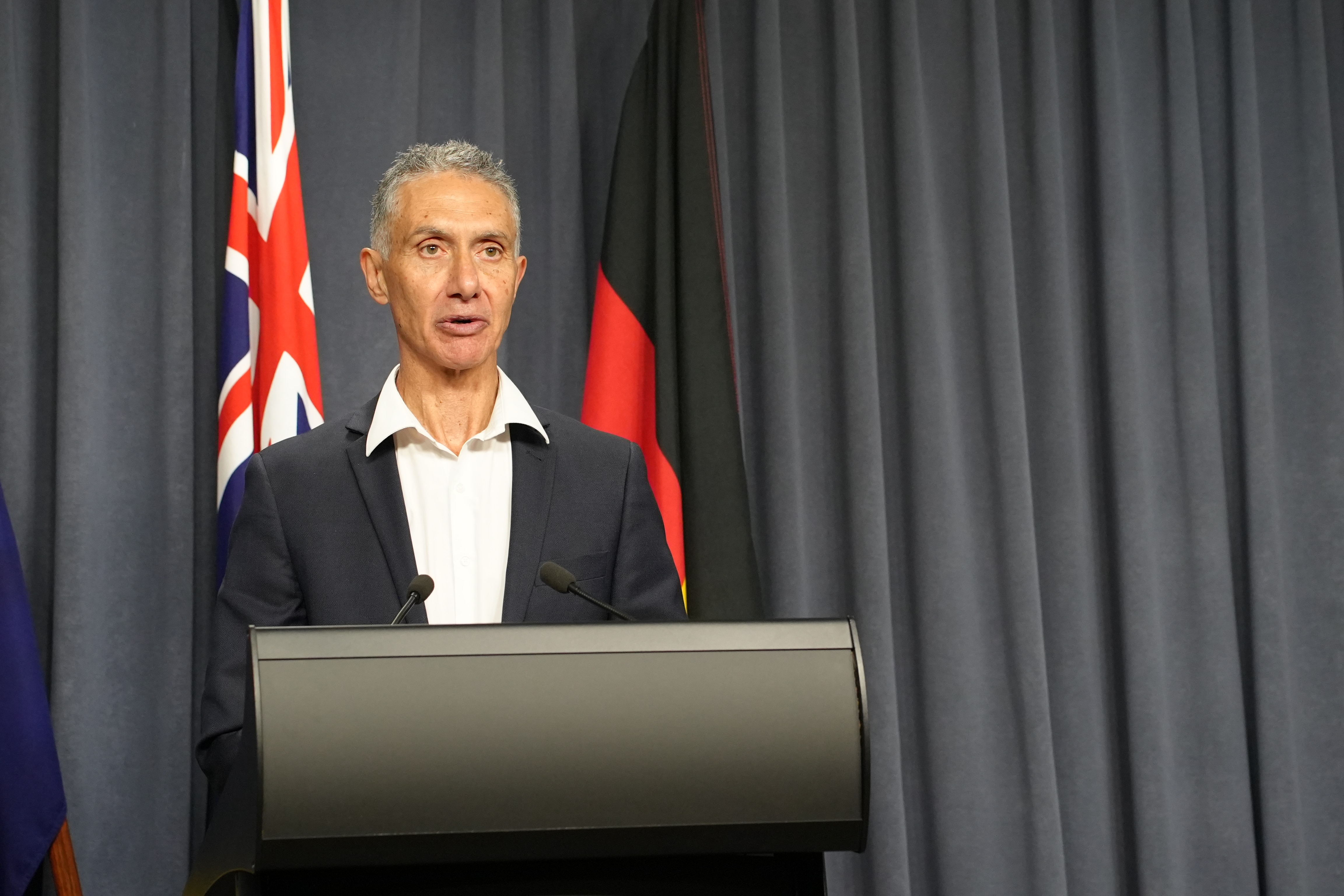 Tertiary Education Minister Tony Buti speaking at a podium at a media conference indoors wearing a suit and white shirt.