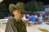 An Aboriginal man in a black striped shirt and wide brimmed hat stands in a outdoor bar area.
