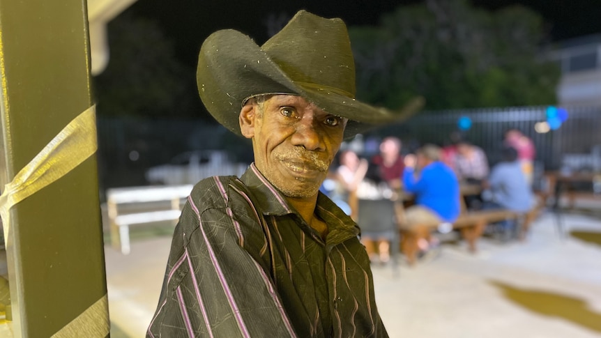 An Aboriginal man in a black striped shirt and wide brimmed hat stands in a outdoor bar area.