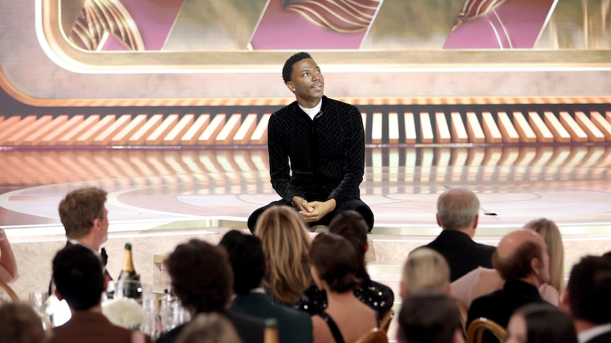 Jerrod Carmichael sits on the steps leading up to a stage, looking to his left as an audience watches him
