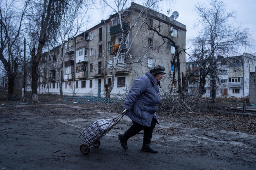 a woman pulls a trolley along a road outside a damaged building in winter