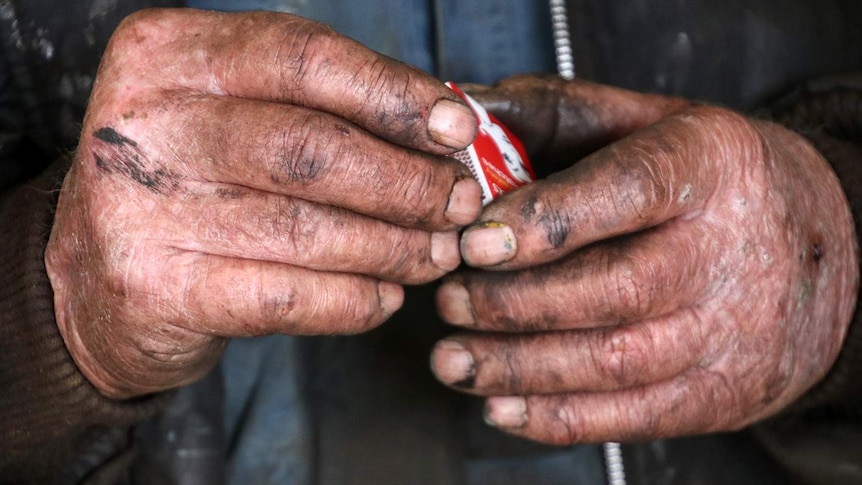 A close up of Bruce's hands holding a bright red matchbox in dirt and oil-stained hands.