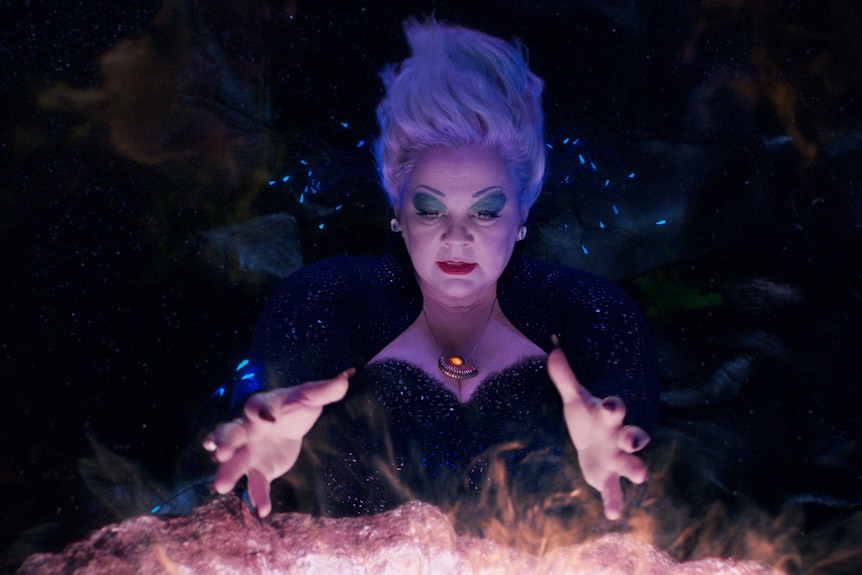 A woman with a silvery beehive hairdo conjures a fiery magic spell. She wears a black bustier and has bold blue eyeshadow on.
