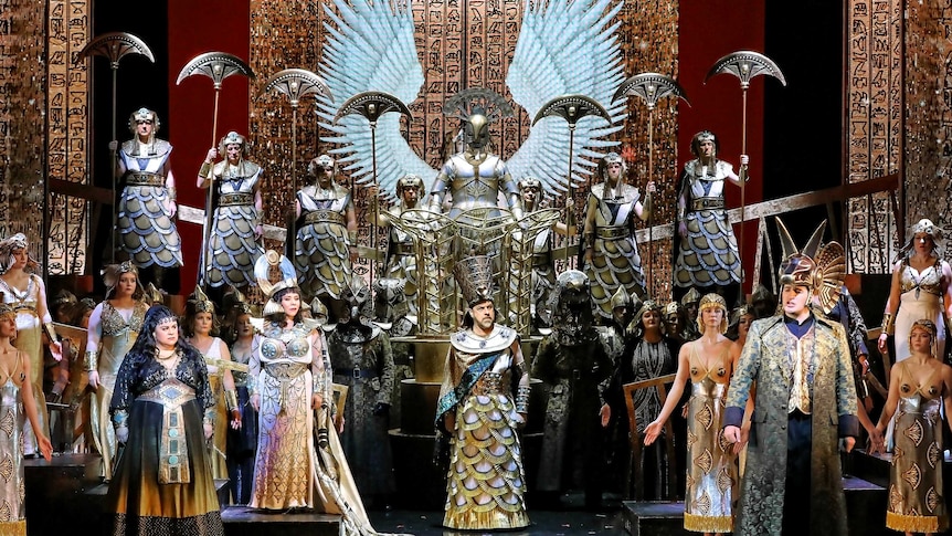A large ensemble of performers in ancient Egyptian costumes stand in front of a digital backdrop of hieroglyphs.