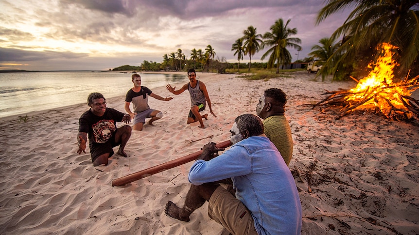 Five men with face paint and didgeridoo sitting on a beach in front of a bonfire